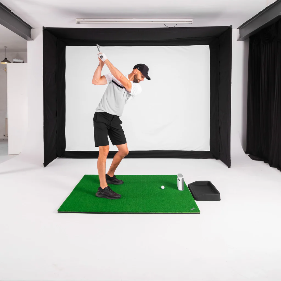 Golf simulator enclosure set up in a bare garage-style room with a golfer mid-swing