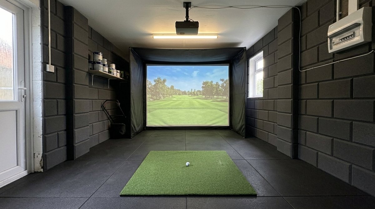 Illustration: golf simulator enclosure, hitting mat and a ceiling-mounted projector in a UK single garage, daylight; black rubber flooring and black foam wall tiles