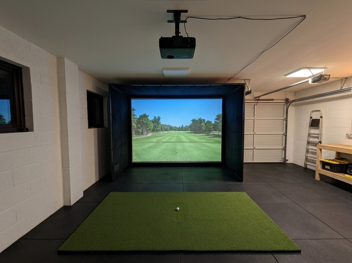 Illustration: golf simulator enclosure, hitting mat and a ceiling-mounted projector in a UK double garage, evening light; black rubber flooring