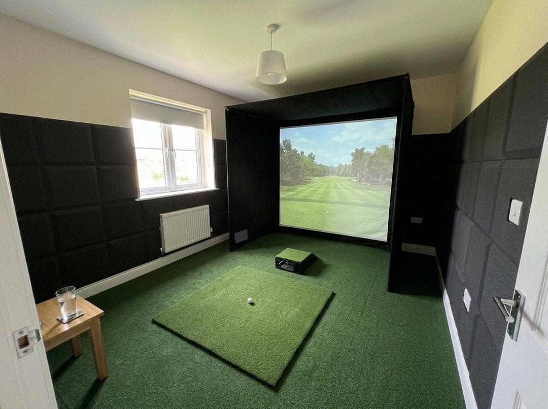 Illustration: golf simulator enclosure, hitting mat and a floor projector case in front of the mat in a UK spare bedroom, daylight; artificial grass flooring and black foam wall tiles