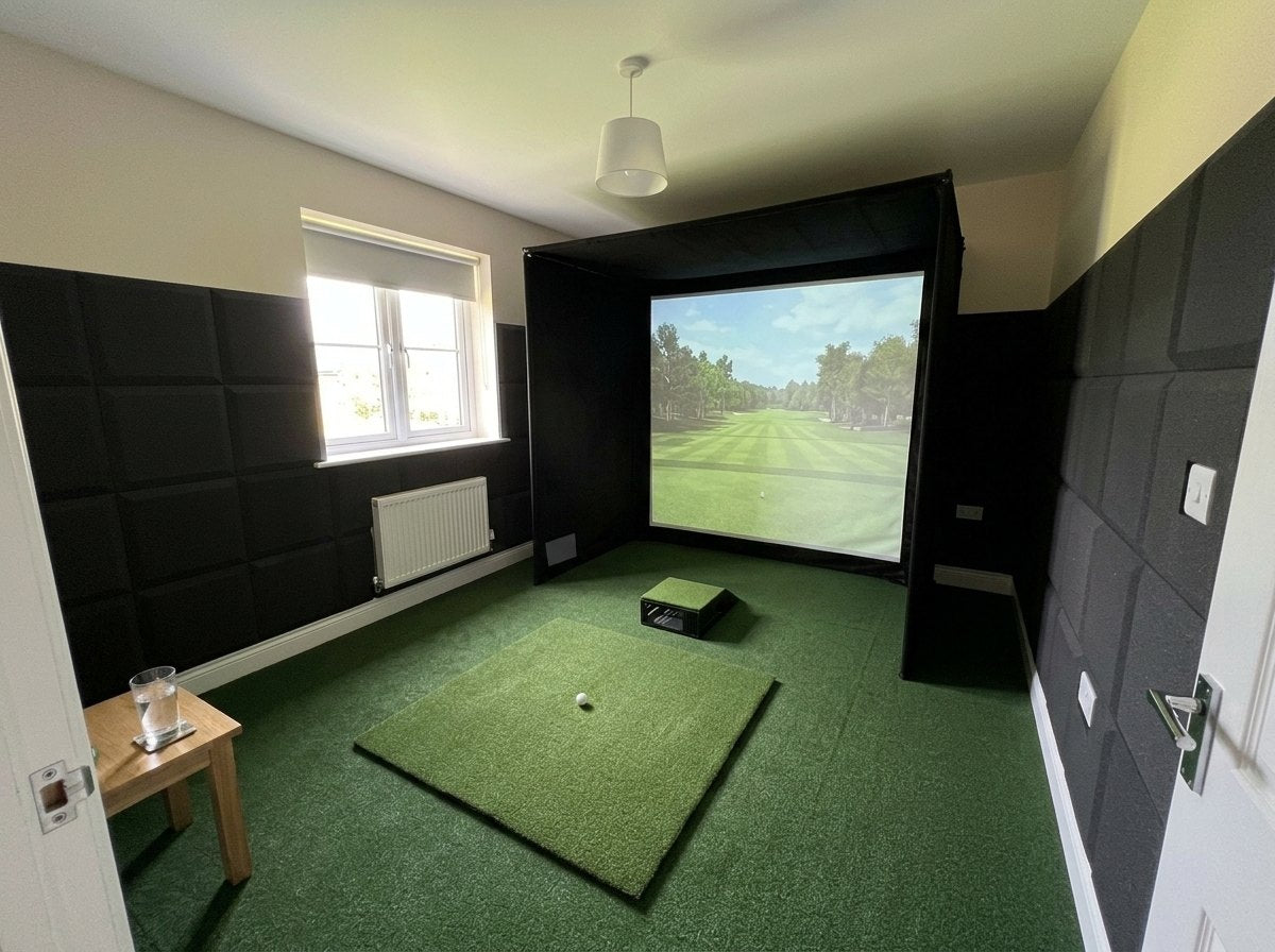 Illustration: golf simulator enclosure, hitting mat and a floor projector case in front of the mat in a UK spare bedroom, daylight; artificial grass flooring and black foam wall tiles