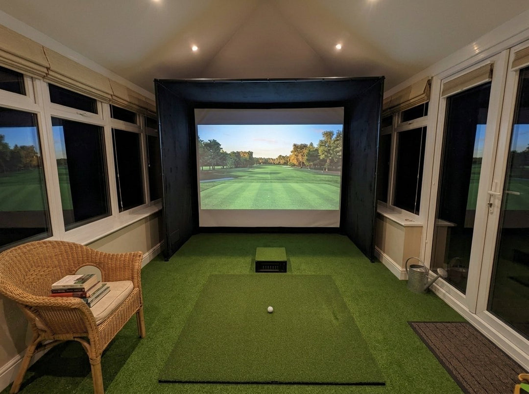 Illustration: golf simulator enclosure, hitting mat and a floor projector case in front of the mat in a UK conservatory, evening light; artificial grass flooring