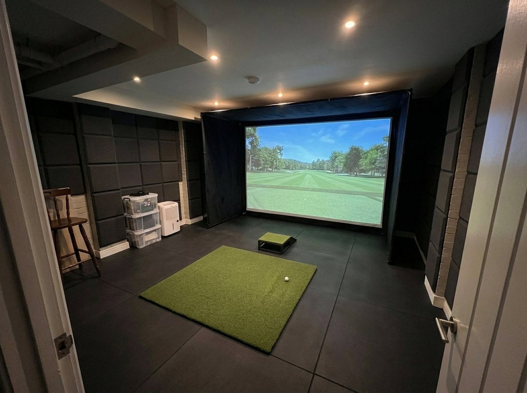 Illustration: golf simulator enclosure, hitting mat and a floor projector case in front of the mat in a UK basement room, evening light; black rubber flooring and black foam wall tiles